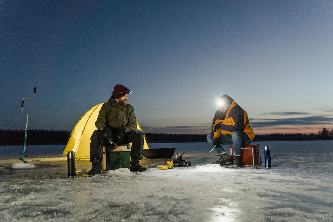 Two men ice fishing by a tent during winter dusk. Adventure on a frozen lake.